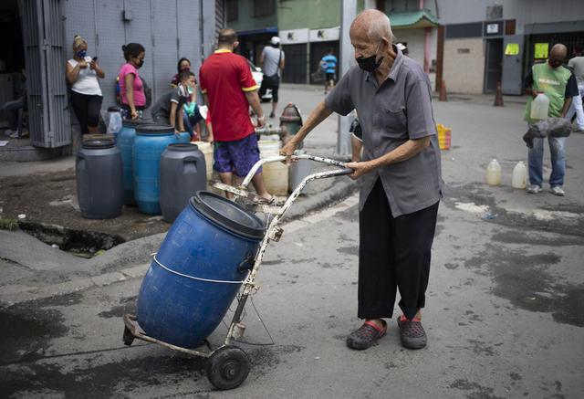 Un hombre, con una máscara facial protectora, empuja una carretilla que sostiene un recipiente lleno de agua que recolectó de un grifo de la calle, en Caracas. (AP Photo/Ariana Cubillos).