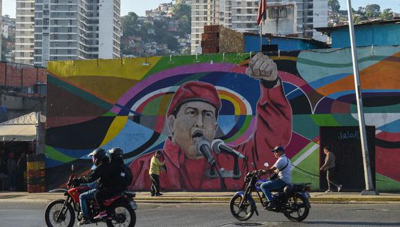 Hombres en motocicletas pasan frente a un mural que representa al difunto presidente de Venezuela Hugo Chávez (1954-2013) en Caracas, Venezuela, el 2 de marzo de 2023. (Foto de Miguel ZAMBRANO / AFP)
