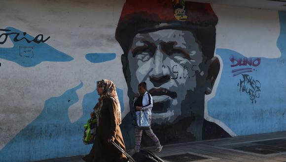 Una mujer que lleva una maleta pasa frente a un mural que representa al difunto presidente de Venezuela, Hugo Chávez (1954-2013), en Caracas, el 2 de marzo de 2023. (Foto de Miguel ZAMBRANO / AFP)