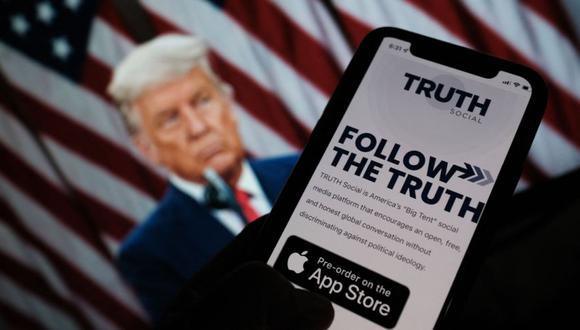 Una persona revisando la tienda de aplicaciones en un teléfono inteligente en busca de "Truth Social", con una foto del expresidente de Estados Unidos, Donald Trump, en la pantalla de una computadora de fondo, en Los Ángeles. (Chris DELMAS / AFP)
