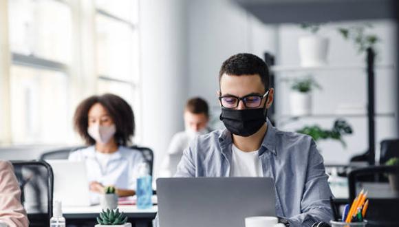 Office center workers are protected from virus outbreak during covid-19 epidemic. Young hipster man in glasses and protective mask works at laptop, with colleagues at workplaces in interior, free space