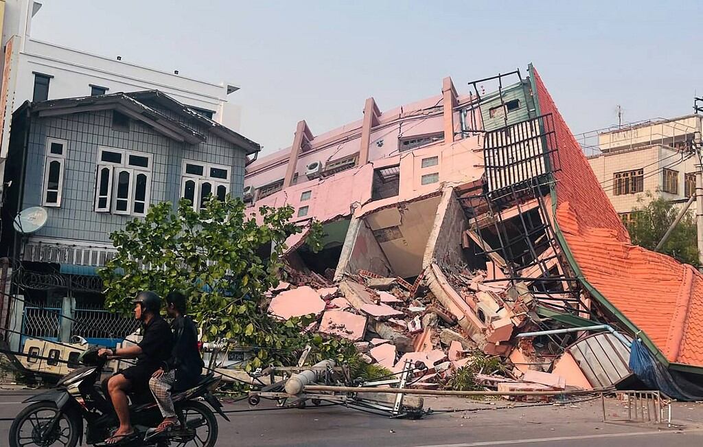 People drive on a motorbike past a collapsed building in Mandalay on March 28, 2025, after an earthquake in central Myanmar. A powerful earthquake rocked central Myanmar on March 28, buckling roads in capital Naypyidaw, damaging buildings and forcing people to flee into the streets in neighbouring Thailand. (Photo by AFP)