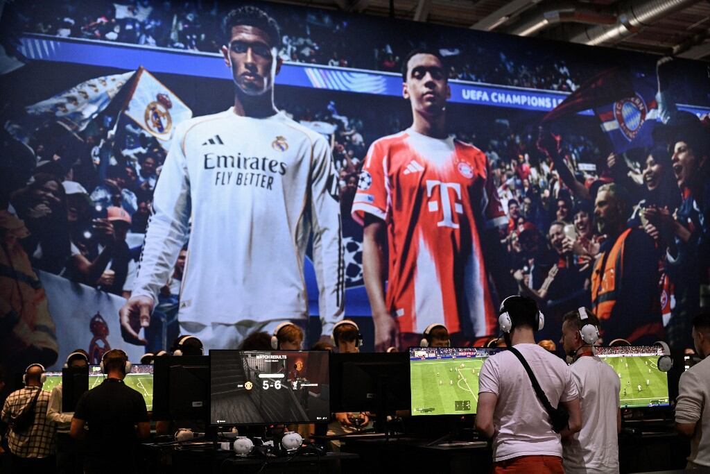 Jugadores disfrutan del videojuego "EA Sports FC 26" frente a una pantalla que muestra imágenes del centrocampista inglés Jude Bellingham (izq.) y del centrocampista alemán del Bayern de Múnich, Jamal Musiala, durante la feria de videojuegos Gamescom, que se celebra en el centro de exposiciones de Colonia, en el oeste de Alemania, el primer día de la feria, el 20 de agosto de 2025. (Foto de INA FASSBENDER / AFP)