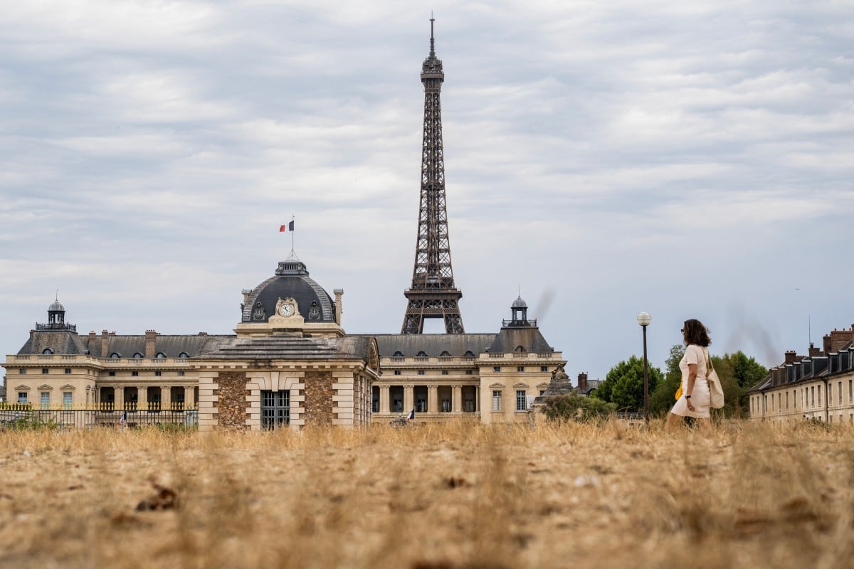 Una mujer camina junto a la Torre Eiffel y el complejo de la École Militaire, con la hierba seca en primer plano, en medio de una ola de calor en París el 13 de julio de 2026. (Foto: Martin LELIEVRE / AFP)