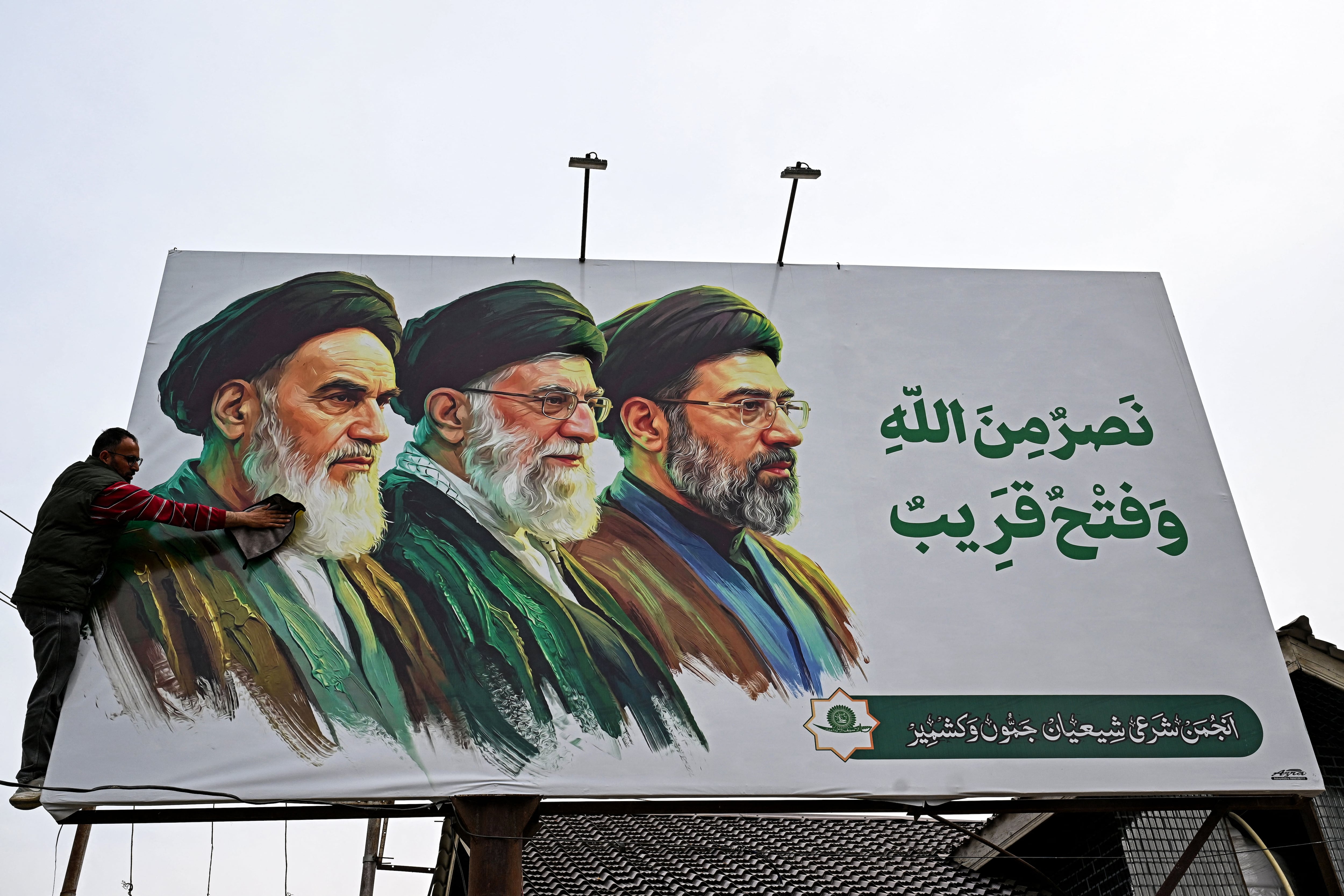Un hombre limpia una valla publicitaria que muestra a los difuntos líderes supremos de Irán, el ayatolá Ruhollah Jomeini (izquierda) y el ayatolá Ali Jamenei (centro), junto al recién elegido líder supremo, Mojtaba Jamenei. (Foto de Tauseef MUSTAFA / AFP).