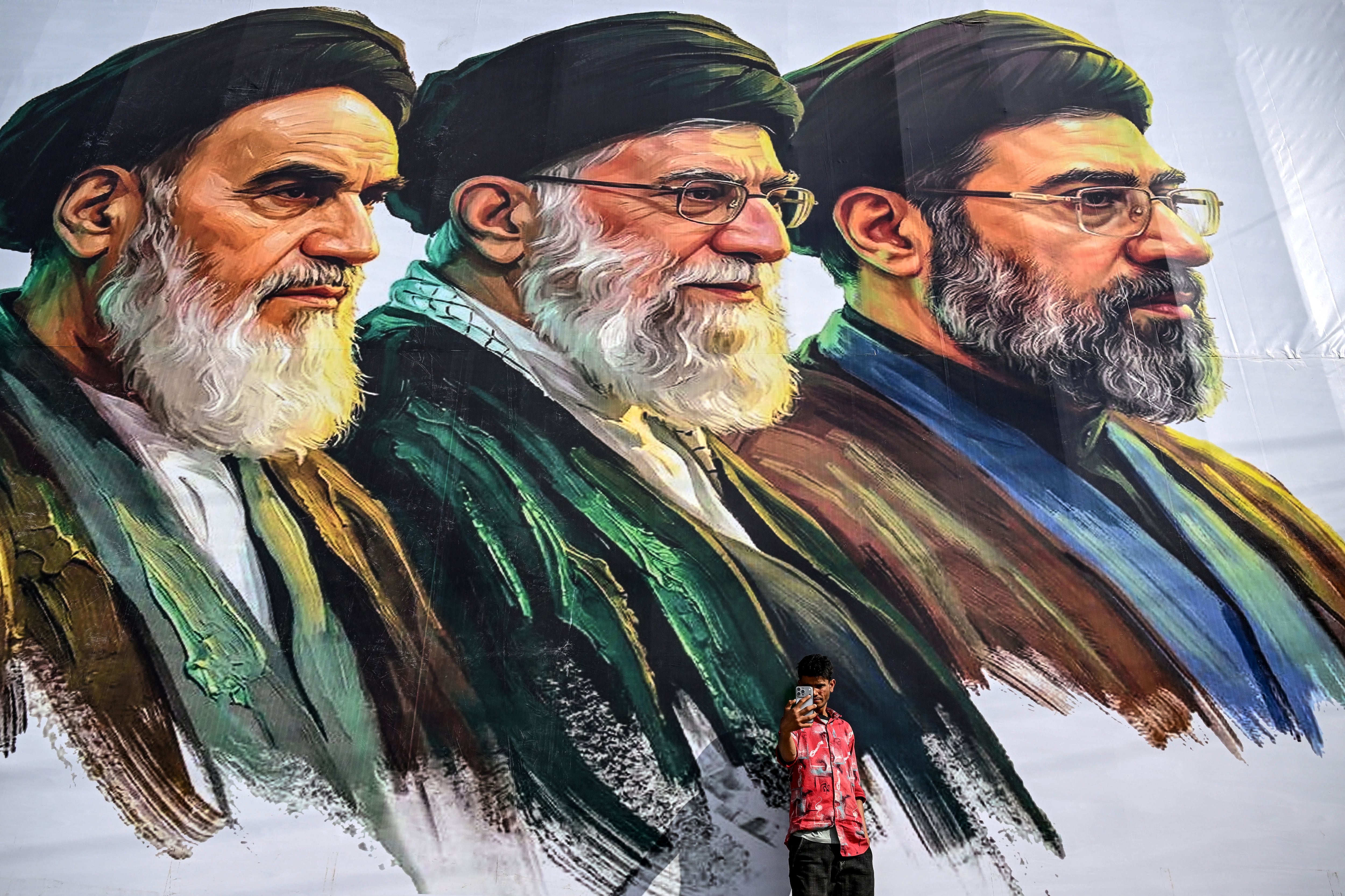 Un hombre se toma una selfie frente a una valla publicitaria que muestra a los difuntos líderes supremos de Irán, el ayatolá Ruhollah Joomeini (izquierda) y el ayatolá Alí Jamenei (centro), junto al recién elegido líder supremo Mojtaba Jamenei. (Foto de Tauseef MUSTAFA / AFP).