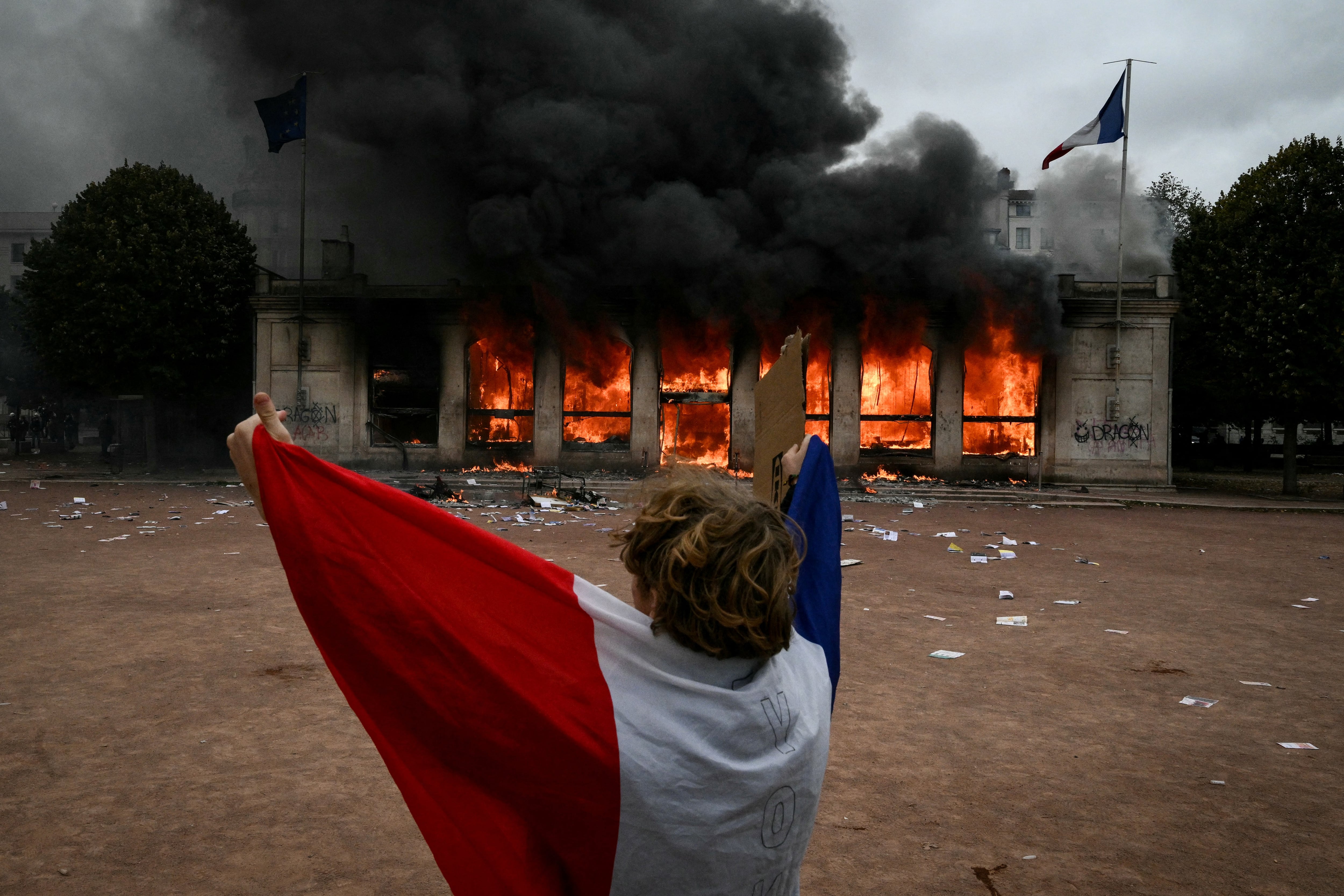 Un manifestante envuelto en la bandera francesa se encuentra frente a la oficina de turismo de Lyon, incendiada durante una protesta contra el alto costo de la vida en el sureste de Francia, el 17 de octubre de 2026. (Foto de Jeff PACHOUD / AFP).