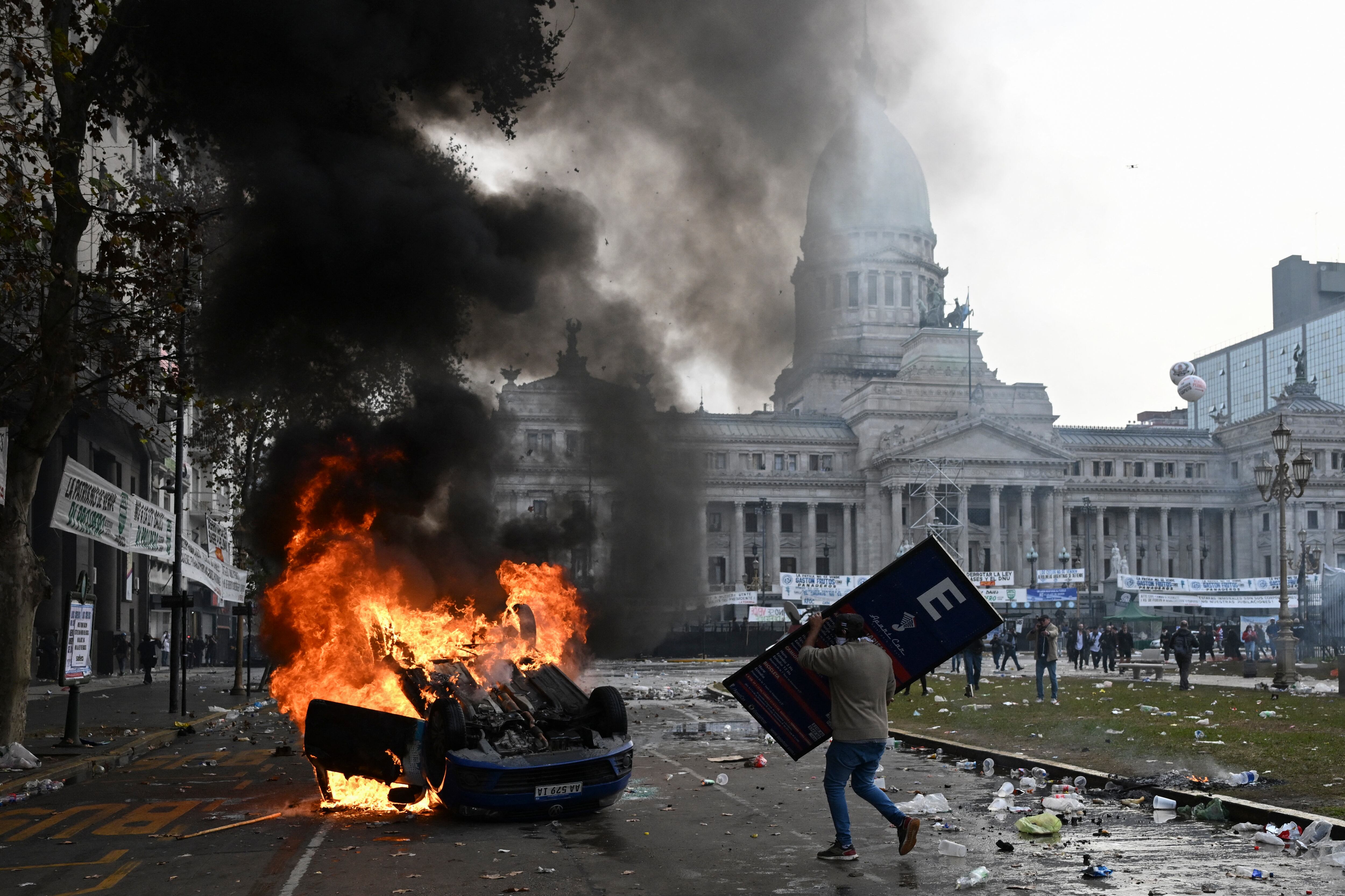 Un manifestante con un cartel se acerca a un automóvil de la estación de radio Cadena 3 incendiado durante una protesta frente al Congreso Nacional en Buenos Aires. (Foto de Luis ROBAYO/AFP).