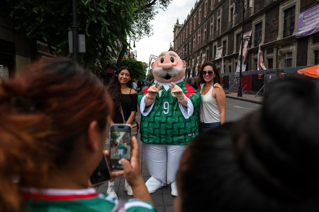 Turistas se toman una foto con un personaje del Dr. Simi vistiendo una camiseta de fútbol mexicano cerca del FIFA Fan Fest en la Plaza del Zócalo, un día antes de la inauguración del torneo de fútbol de la Copa Mundial de la FIFA 2026 en la Ciudad de México el 10 de junio de 2026. (Foto de Gerardo Luna / AFP)