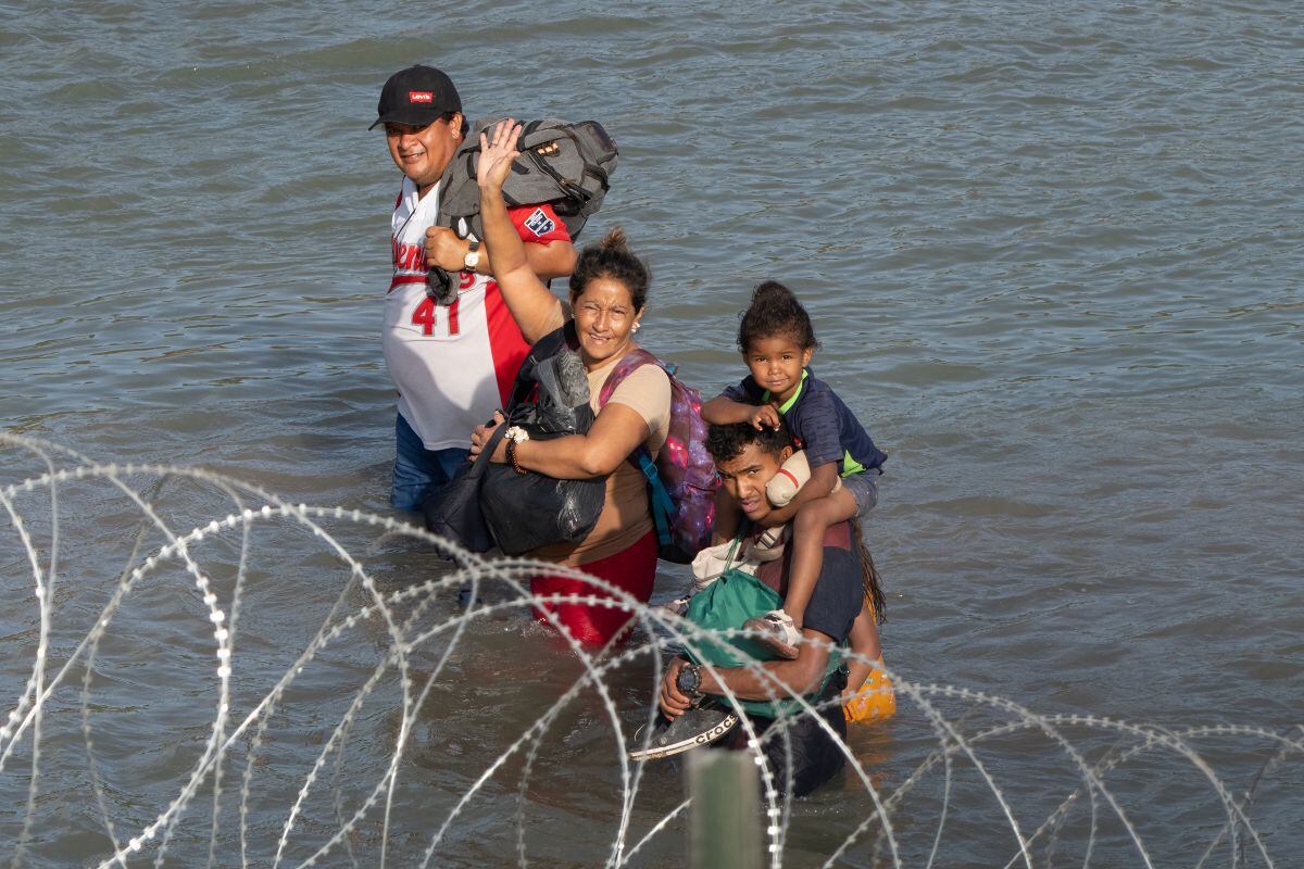 Los migrantes saludan mientras caminan cerca de alambre de púas en el agua a lo largo de la frontera del Río Grande con México en Eagle Pass, Texas, el 16 de julio de 2023 (Foto: Suzanne Cordeiro / AFP)