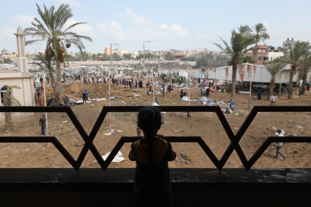 Una niña desplazada observa desde el balcón de una casa cómo palestinos inspeccionan los daños en torno a un cráter provocado por un impacto israelí en un campamento para desplazados internos en Deir el-Balah, en el centro de la Franja de Gaza, el 21 de agosto de 2025. Israel atacó la ciudad de Gaza y sus alrededores durante la noche, según informaron los residentes el 20 de agosto, mientras el ejército anunciaba los primeros pasos en su ofensiva para capturar el último bastión importante de Hamás. (Foto de Eyad BABA / AFP)