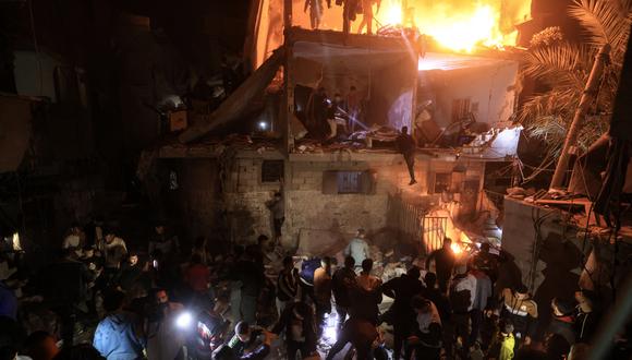 La gente observa mientras otros buscan víctimas entre los escombros de un edificio en llamas, tras un ataque de Israel en Rafah, en el sur de la Franja de Gaza, el 6 de diciembre de 2023. (Foto de Mahmud HAMS / AFP).