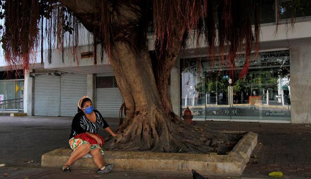 Una mujer descansa bajo un árbol frente a locales comerciales cerrados en Cartagena (Colombia). (EFE/ Ricardo Maldonado Rozo).